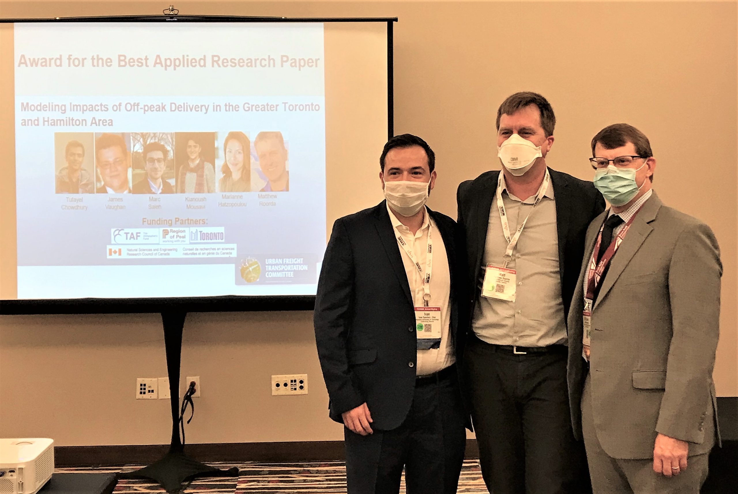 Three men wearing masks stand in front of projection of winning paper